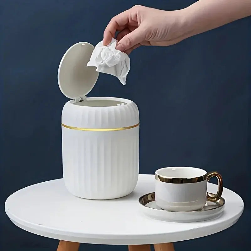 Mini desktop trash can with lid, white ribbed with a gold band, on a round white table beside a white and gold teacup