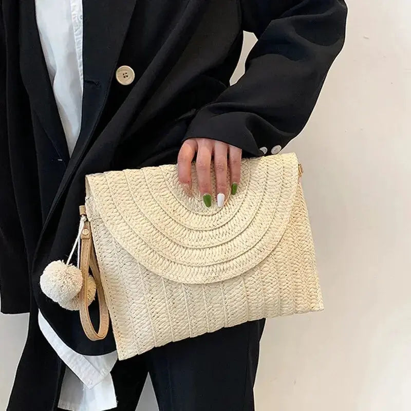 Handmade straw crossbody bag with an envelope-style curved flap, beige woven texture and a pom-pom charm