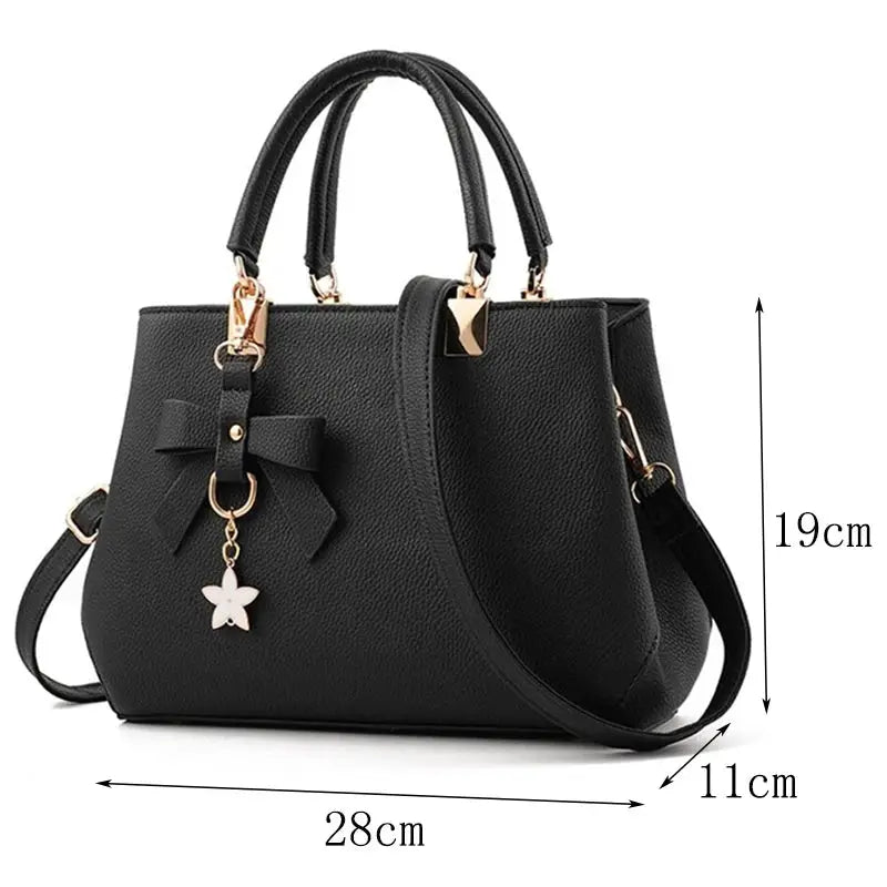 Black pebbled leather handbag with dual top handles, detachable shoulder strap, gold hardware, flower bow charm.