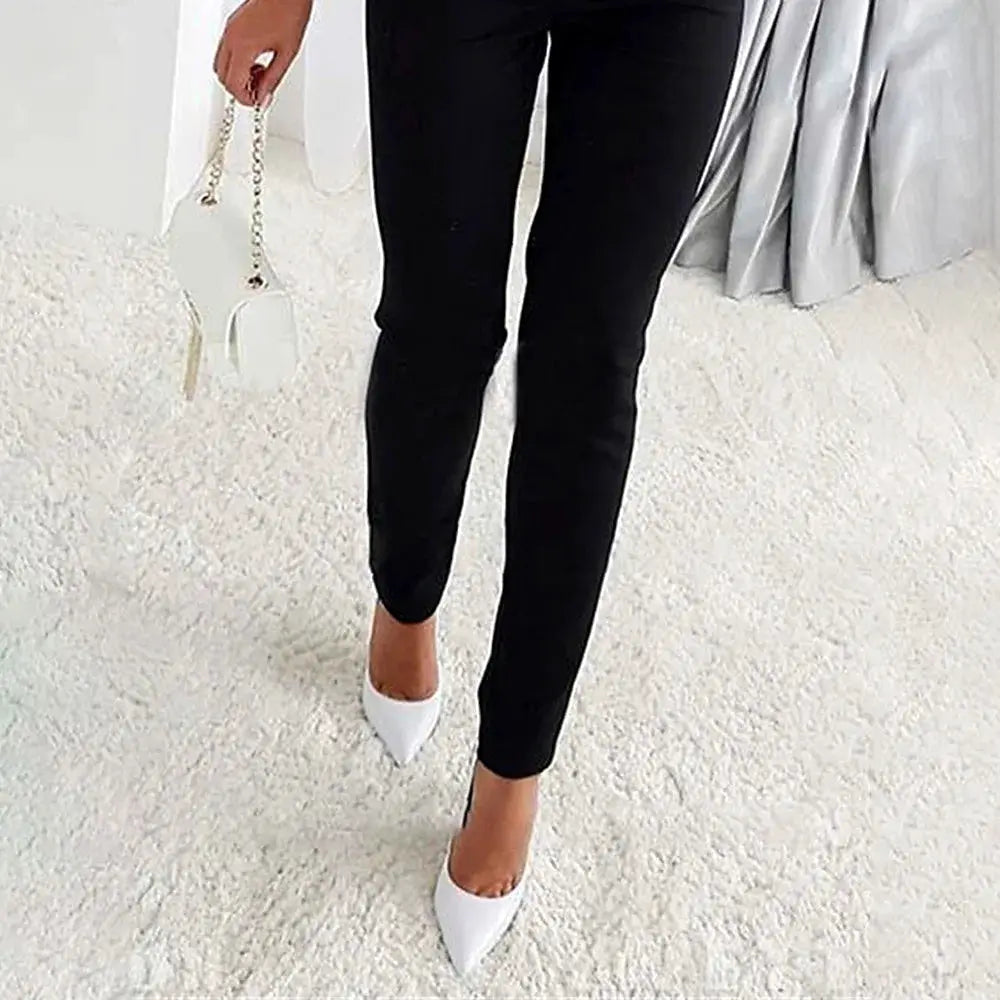 Black women's one-piece jumpsuit with slim fit and high waist, paired with white pointed-toe heels and a white handbag.