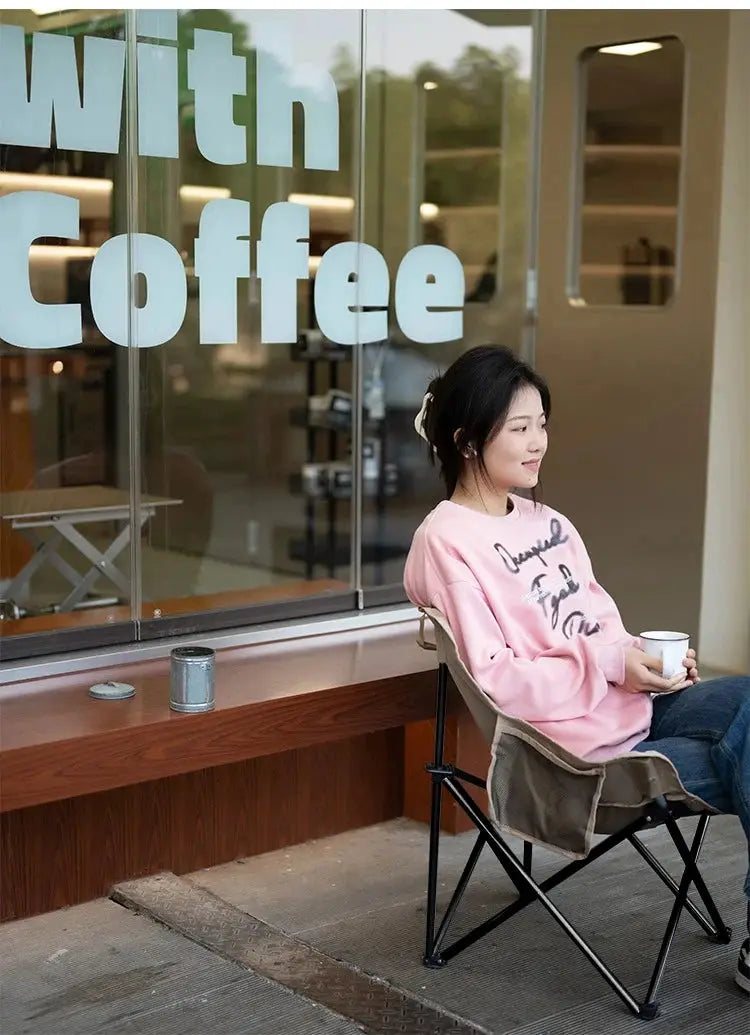 Outdoor folding moon chair beige with black frame beside a cafe window, as a woman in a pink hoodie relaxes with a cup.