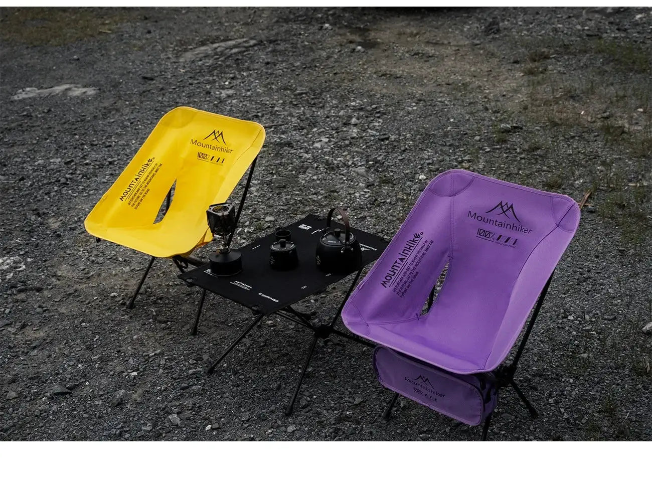 Two foldable camping chairs in yellow and purple with a table between them on rocky ground.