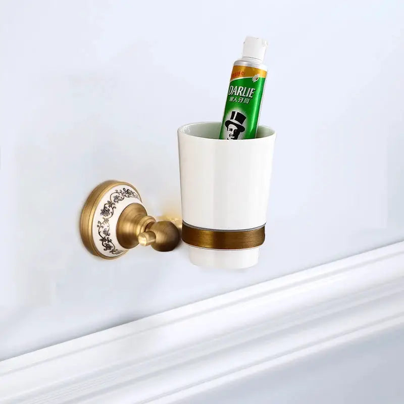 Antique brushed solid brass wall-mounted toothbrush holder with white porcelain cup and decorative backplate.