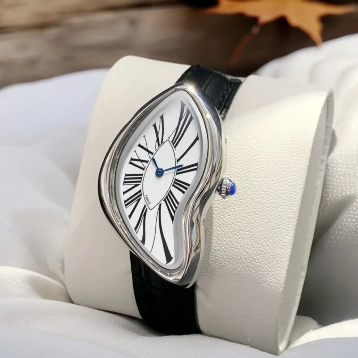 Wristwatch with a warped silver case, white Roman-numeral dial, blue hands, and black leather strap on a white pillow
