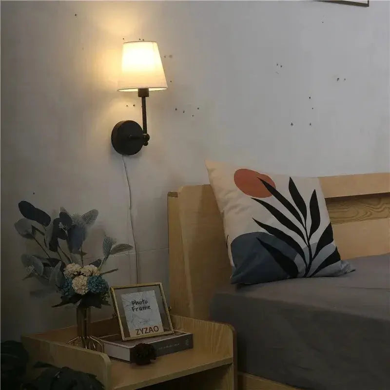 American Retro wall lamp with cream fabric shade on a black round backplate, mounted beside bed