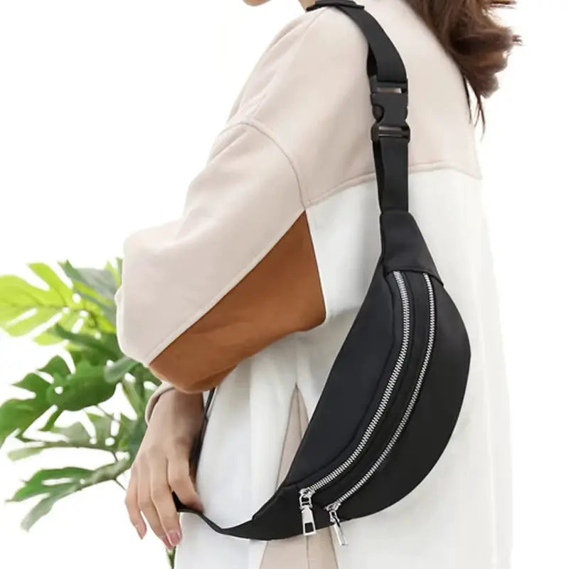 Black sling fanny pack waist bag with two parallel zippers and adjustable strap, worn as crossbody by model