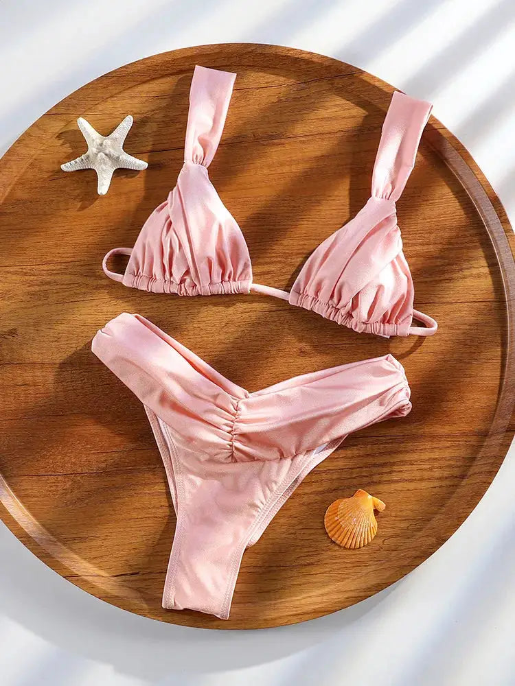 Two-piece bikini set in pastel pink with ruched triangle top and ruched bottom on a wooden tray
