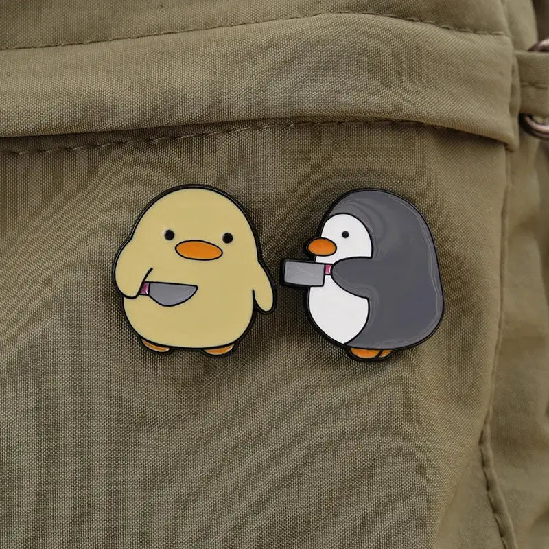Enamel pin set: pair of cartoon penguin pins on khaki backpack, yellow chick and gray penguin with orange beaks