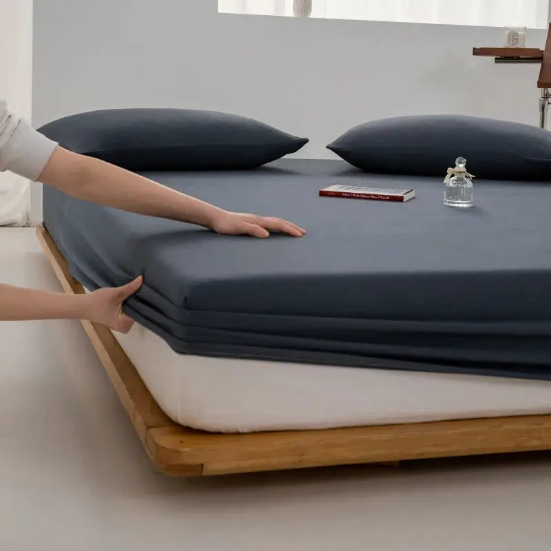 Soft cotton fitted sheet with elastic band in navy blue on a wooden bed frame, fits multiple mattress sizes.