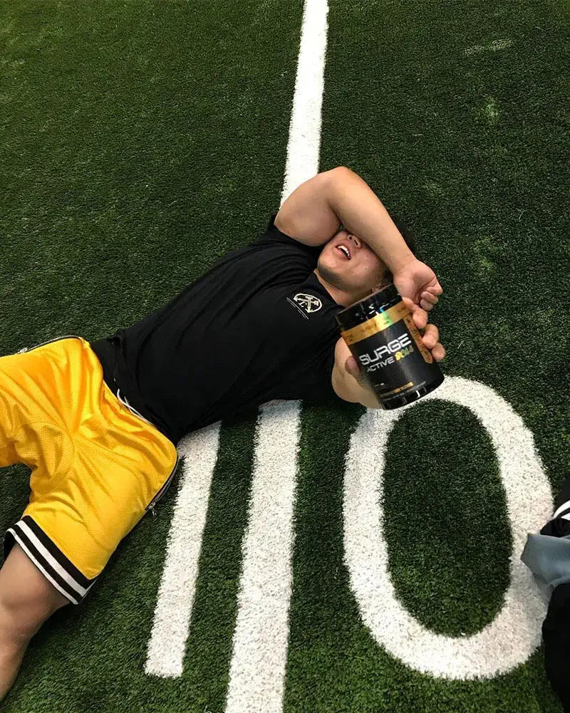 Men's basketball shorts in bright yellow with black trim, worn on turf by a person holding a supplement tub.