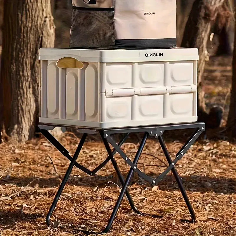 Portable folding camping table with black metal X-frame legs and a white plastic storage box on top for outdoor use