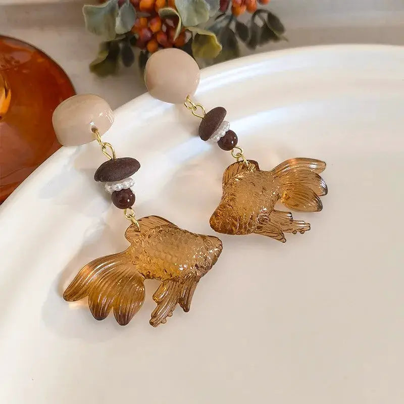 Goldfish pendant earrings with translucent amber fish, cream round tops, and brown bead accents on gold-tone fixtures