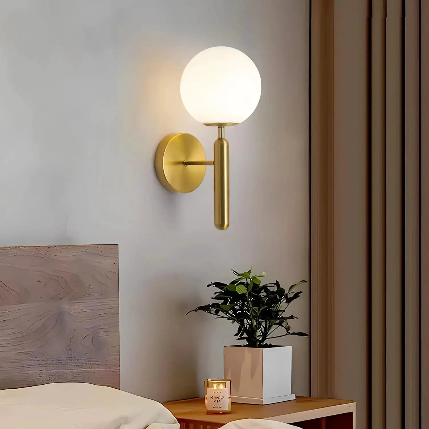 Modern gold wall lamp with frosted glass globe and cylindrical stem mounted beside a bedside table.