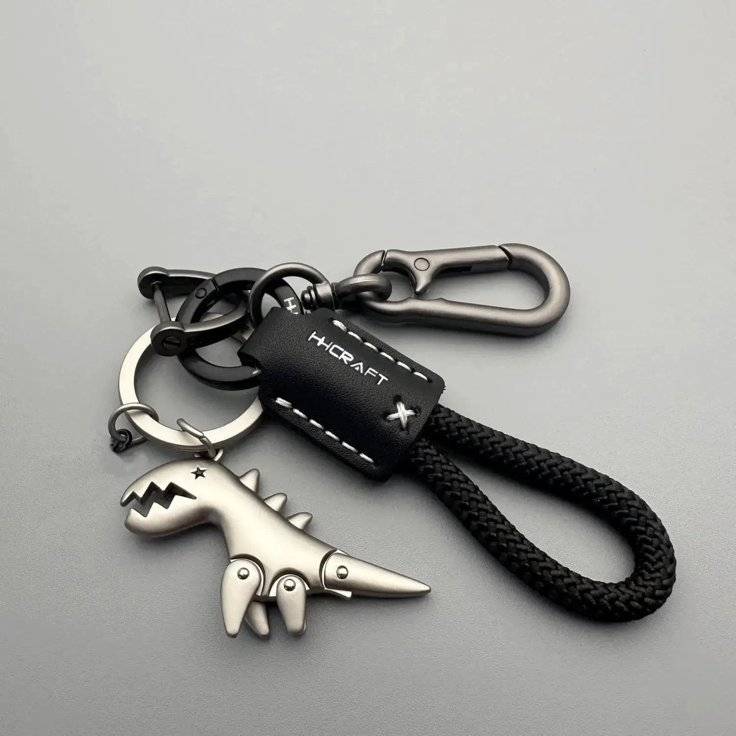 Keychain with black braided rope, leather tag marked HCRAFT, and metallic dinosaur charm on gunmetal hardware.