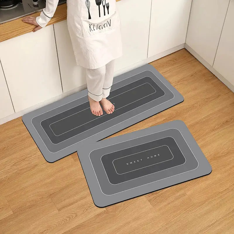 Two gray diatomite kitchen floor mats, waterproof and anti-skid, with border design; a barefoot person stands on one.
