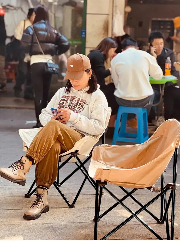 Outdoor folding moon chairs; two tan/beige chairs with black frames, person seated and using a phone on a street.