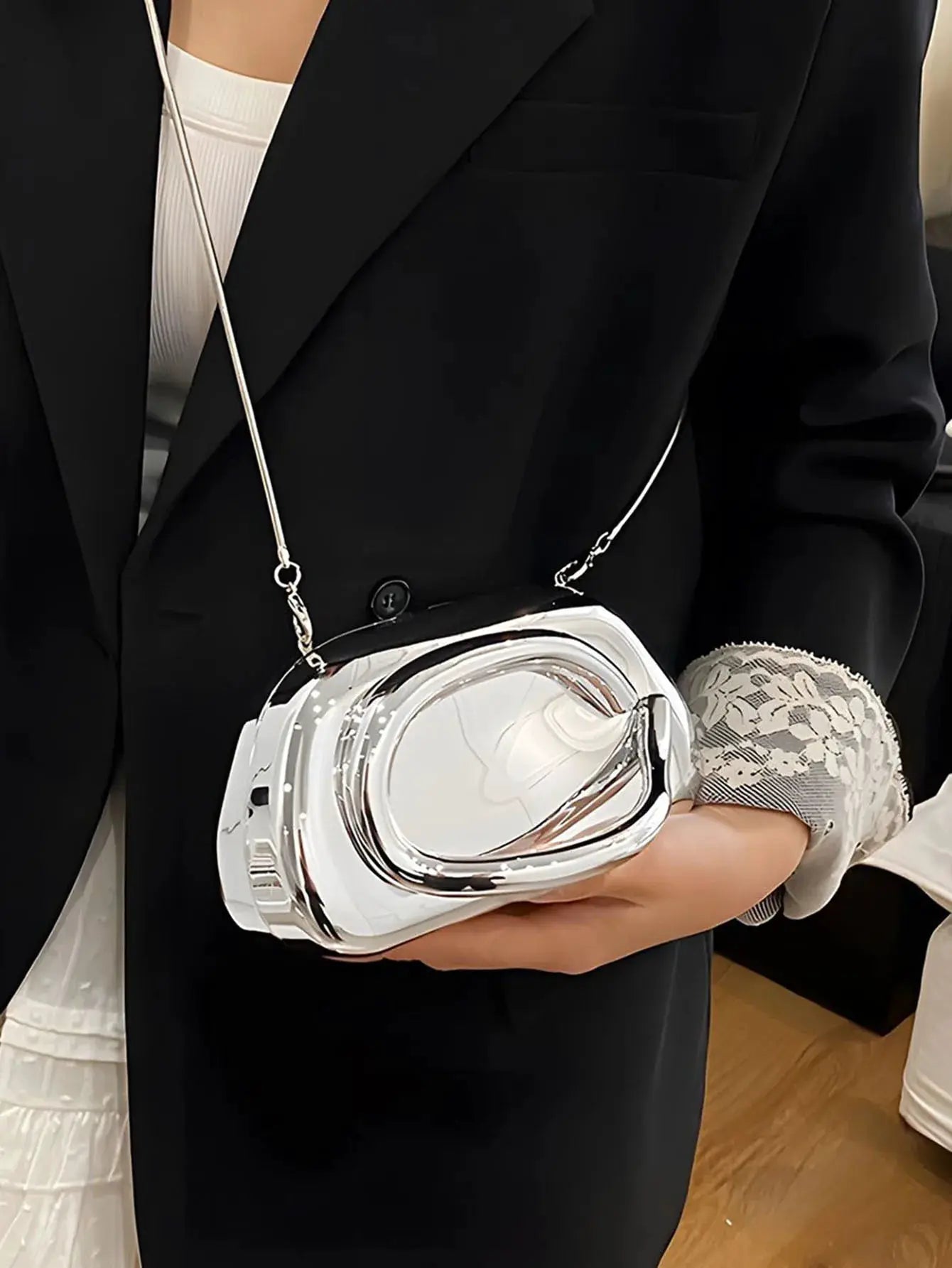 Acrylic box crossbody bag in metallic white with a silver strap, held by a person in a black blazer.