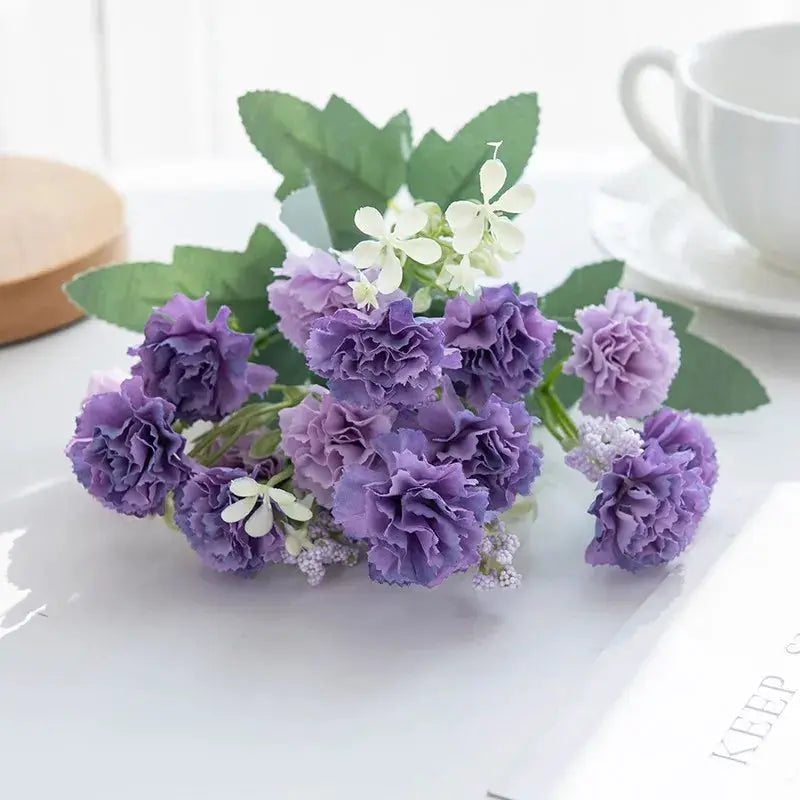 Purple artificial carnations bouquet by Simples Market with white filler blossoms and green leaves
