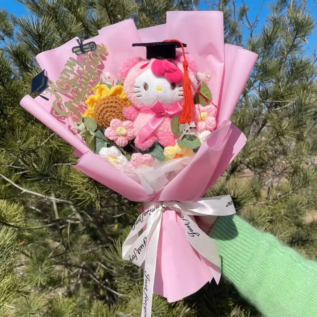 Stuffed plants bouquet with pink Hello Kitty plush wearing a graduation cap, surrounded by crochet flowers.