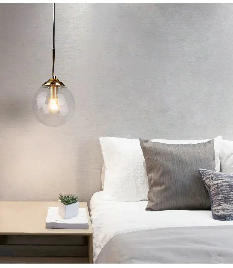 Modern luxury metal glass pendant lamp with a clear globe and brass accents over a white bed.
