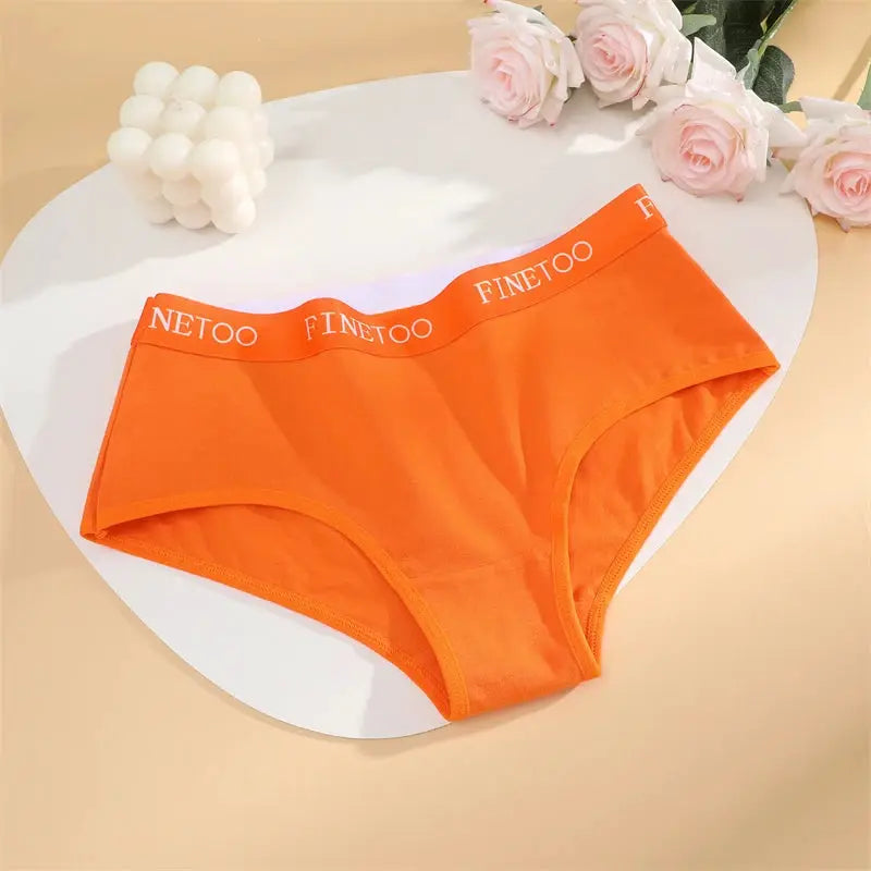 Orange cotton women's briefs with white FINETOO waistband, displayed on a round white table beside pink roses