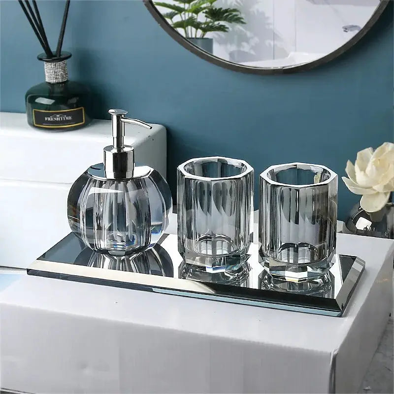 Clear crystal glass bathroom set with lotion pump, toothbrush holder and cup on a mirrored tray.