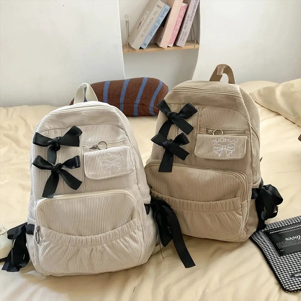Two beige corduroy backpacks with black bow accents and front pockets