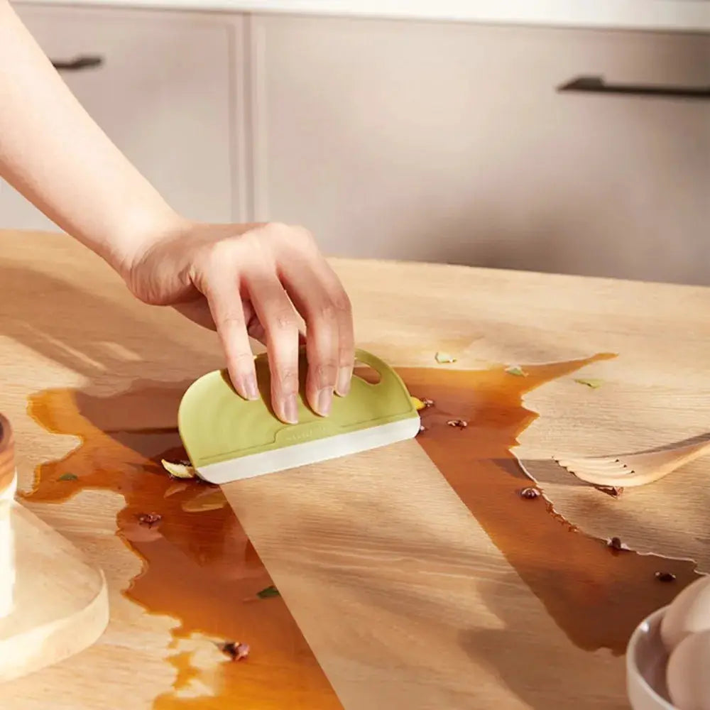 Green silicone handheld kitchen squeegee cleaning a spill on a wooden countertop; non-scratch design