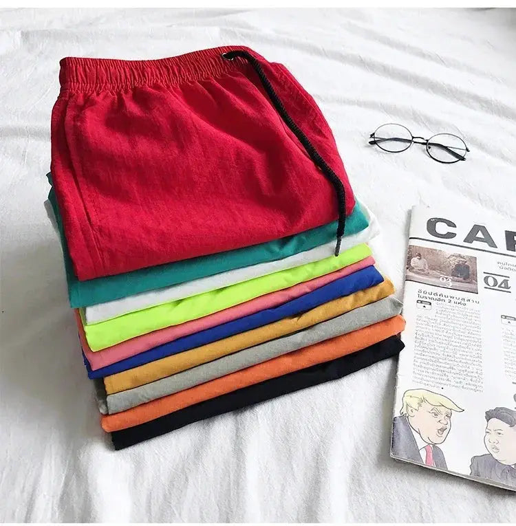 Simples Market men's swim shorts stack; red pair on top with drawstring among colorful beach board shorts.