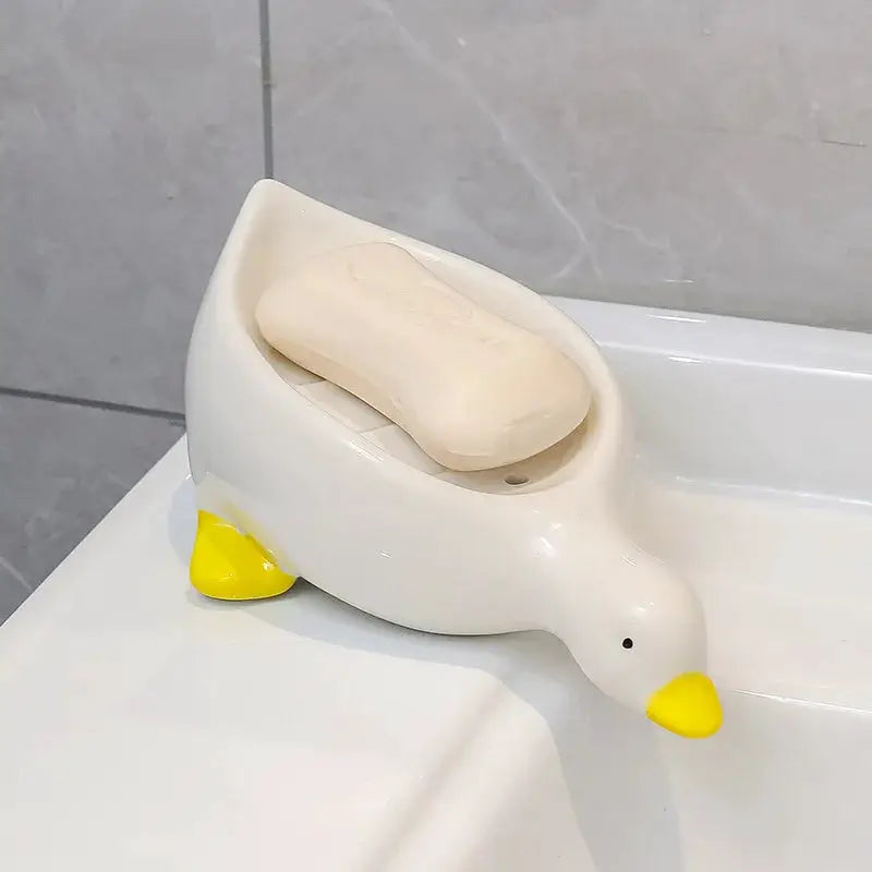 Duck-shaped plastic soap rack, white body with yellow beak and feet, on bathroom tub edge