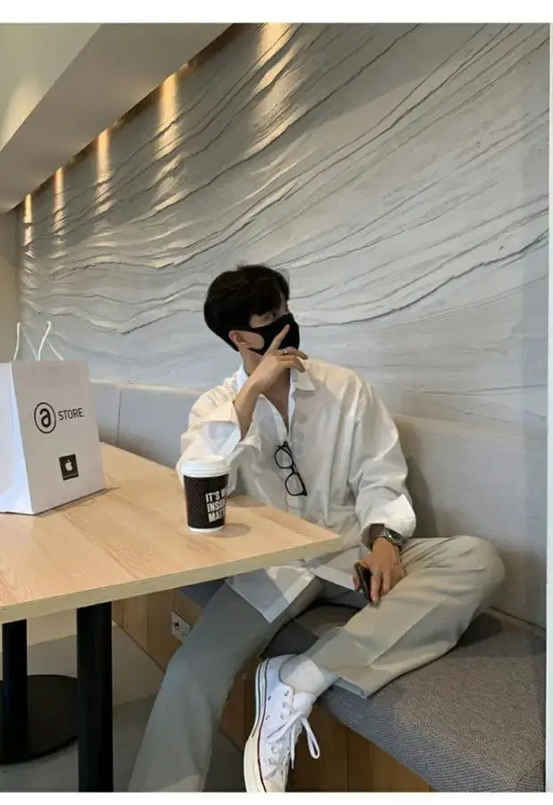 Men's white long-sleeve shirt with beige trousers and white sneakers, seated in a cafe.