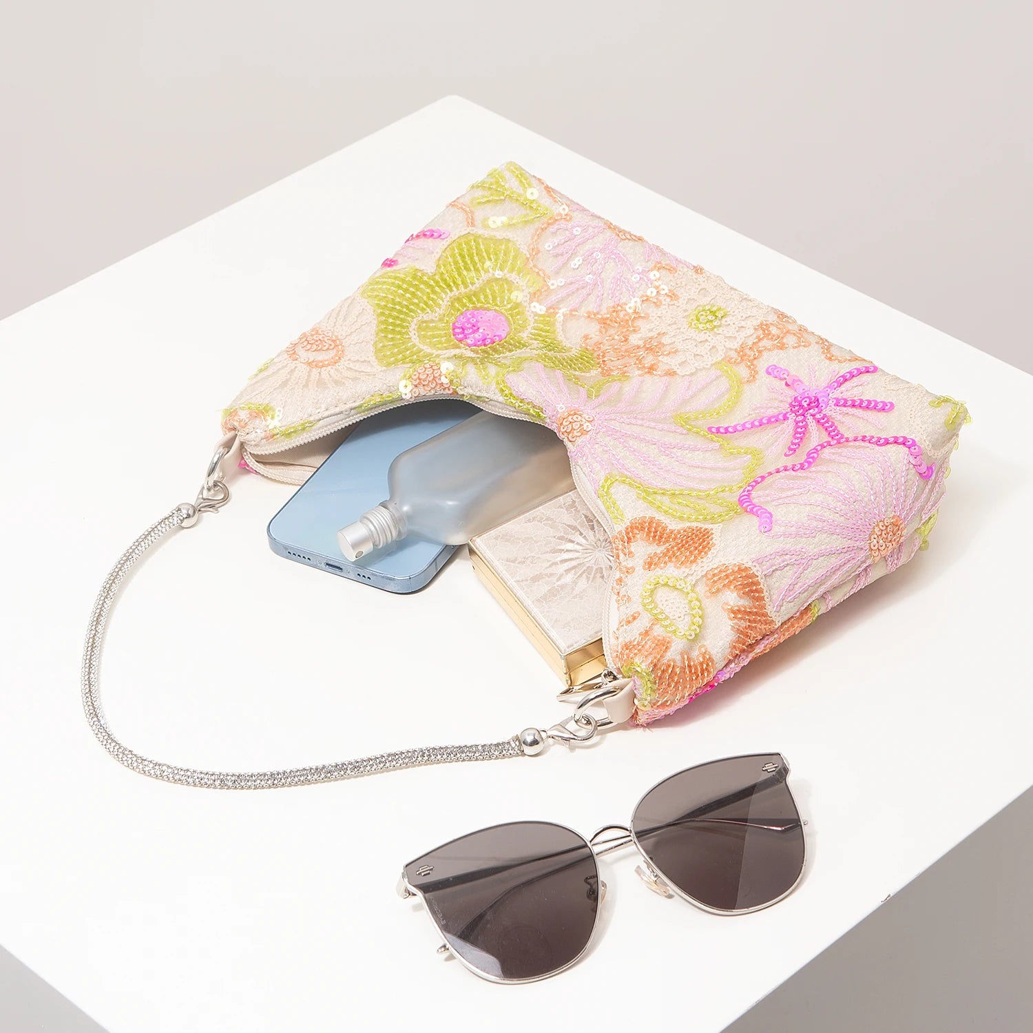 Shoulder bag with pastel pink floral embroidery and sequins, silver chain strap; phone and sunglasses nearby.