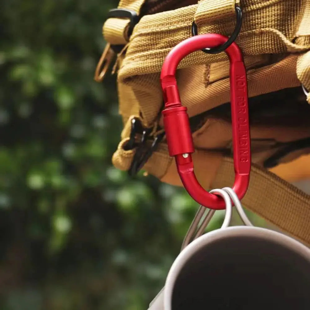 Red aluminium locking D-shaped carabiner on tan backpack strap, with a mug attached.