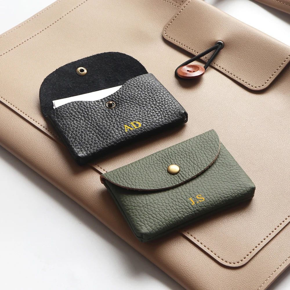 Two pebble-grain leather card holders in black and olive with engraved initials on a beige surface