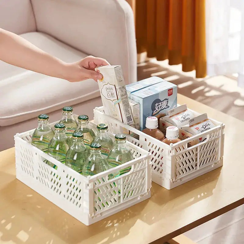 Two white plastic foldable storage crates with lattice sides on a table, holding bottles and boxes