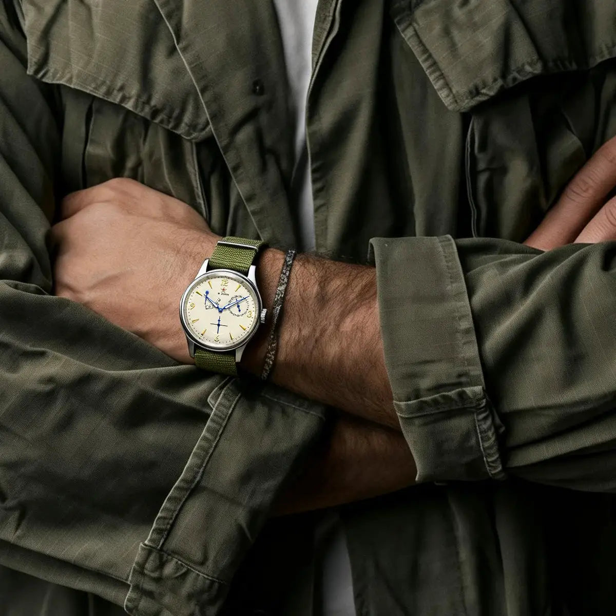 Pilot-style watch with cream dial, blue hands and gold markers on a stainless steel case and olive-green strap.