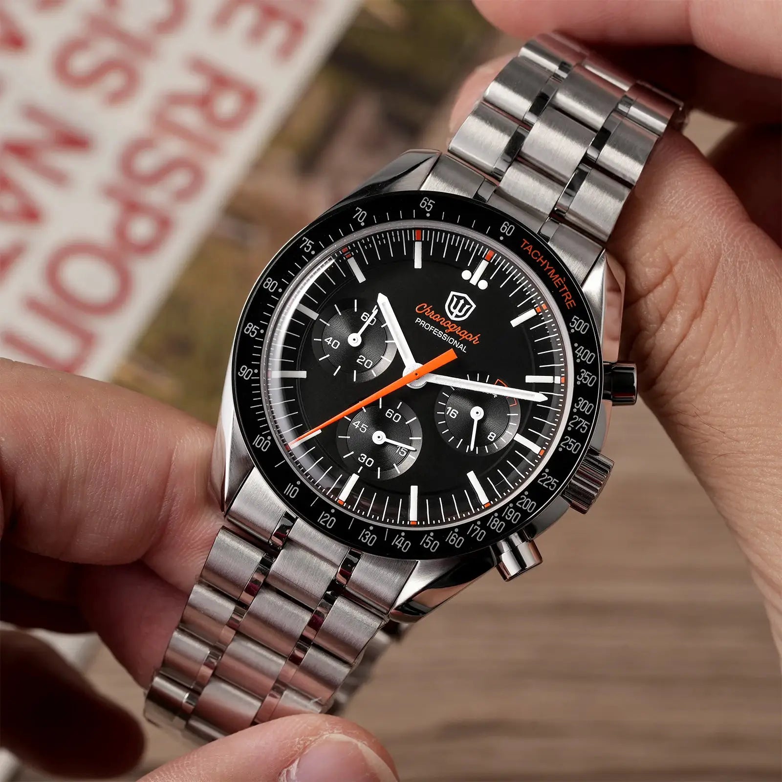 Chronograph watch with stainless steel bracelet, black dial, orange hands and tachymeter bezel.