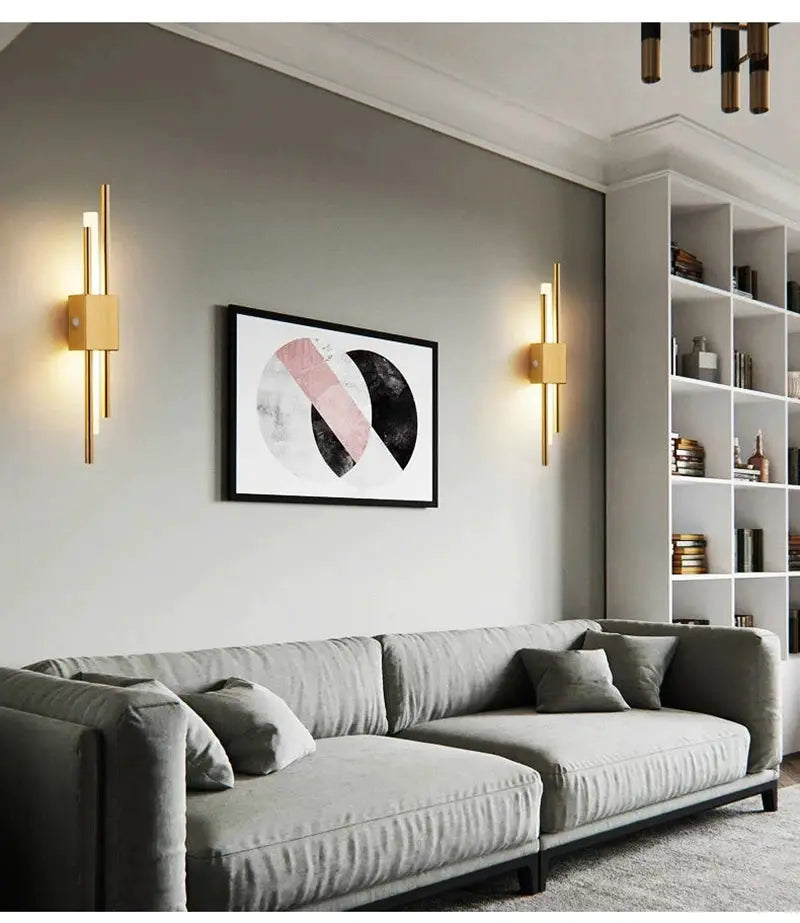 Two Nordic LED wall lamps in gold finish mounted on a gray wall beside a sofa and abstract art.