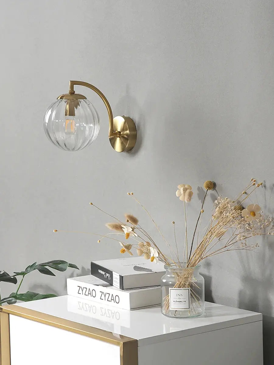 Modern LED wall lamp with a gold brass curved arm and clear glass globe shade on a gray wall.