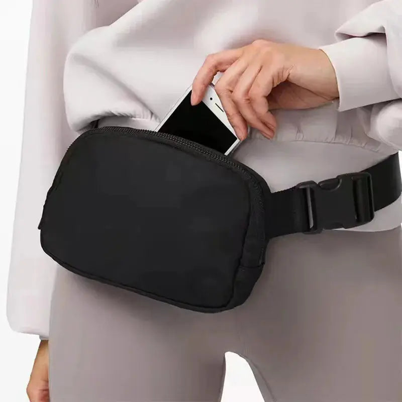 Black waist bag with zipper and adjustable strap worn at the hip; phone tucked in.