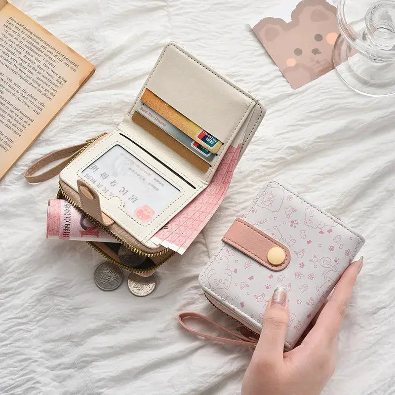 Two cat-themed wallets: a cream zip wallet with card slots and ID window, and a pink patterned compact wallet with strap