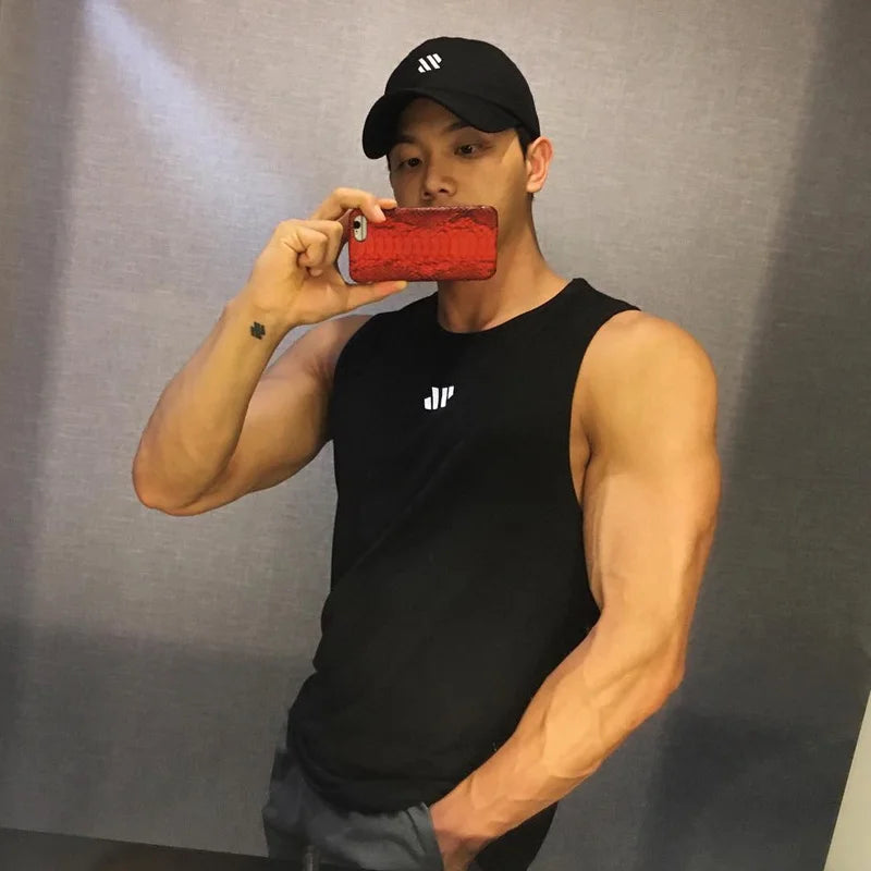 Men's black stringer bodybuilding tank top with small chest logo; man in cap taking a mirror selfie.