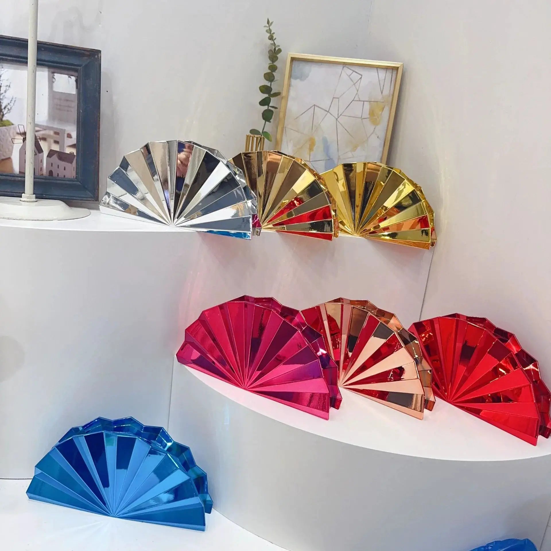 Decorative foil foldable fans in blue, pink, and gold arranged on a white shelf beside a blue-framed mirror