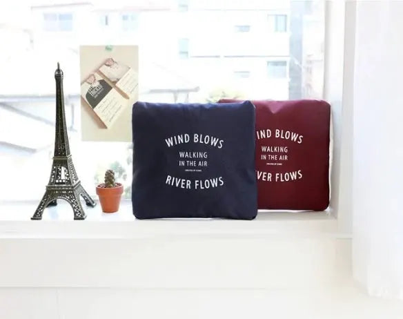 Two square cushions in navy and burgundy with white printed text on a windowsill beside a small Eiffel Tower statue and a plant.