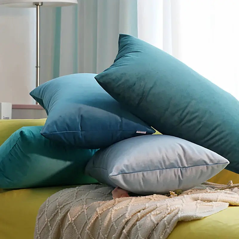 Five soft velvet cushions in teal and blue stacked on a yellow sofa with a knit throw