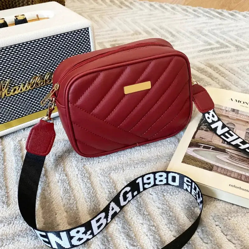 Red quilted PU shoulder bag with a gold plaque and a black strap with white lettering.