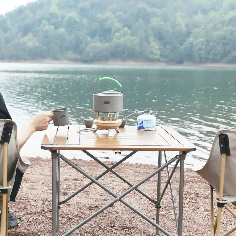 BRS201A camping gas stove with green handle and piezo ignition on a wooden lakeside table; mug nearby.