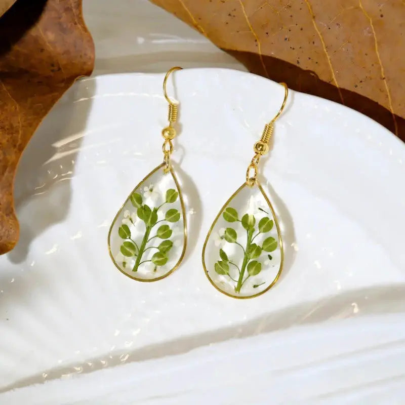 Pair of teardrop epoxy resin earrings with pressed green leaves and tiny white flowers in a gold frame.