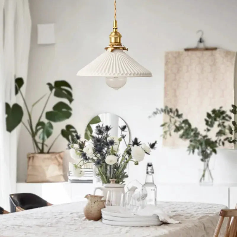 Pendant lamp by Simples Market, white pleated shade with brass hardware in a dining room.
