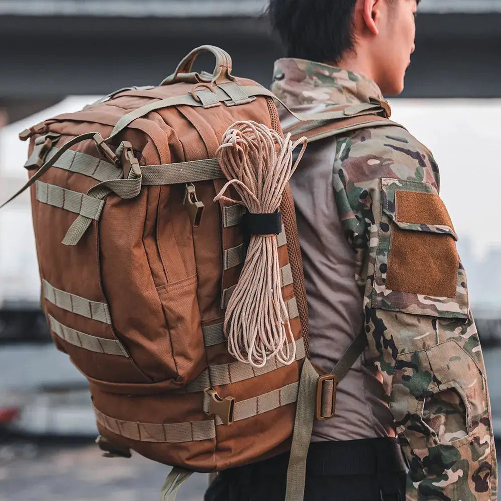 Backpack with rope coil and tactical straps, worn by a person in camouflage jacket in an outdoor setting.